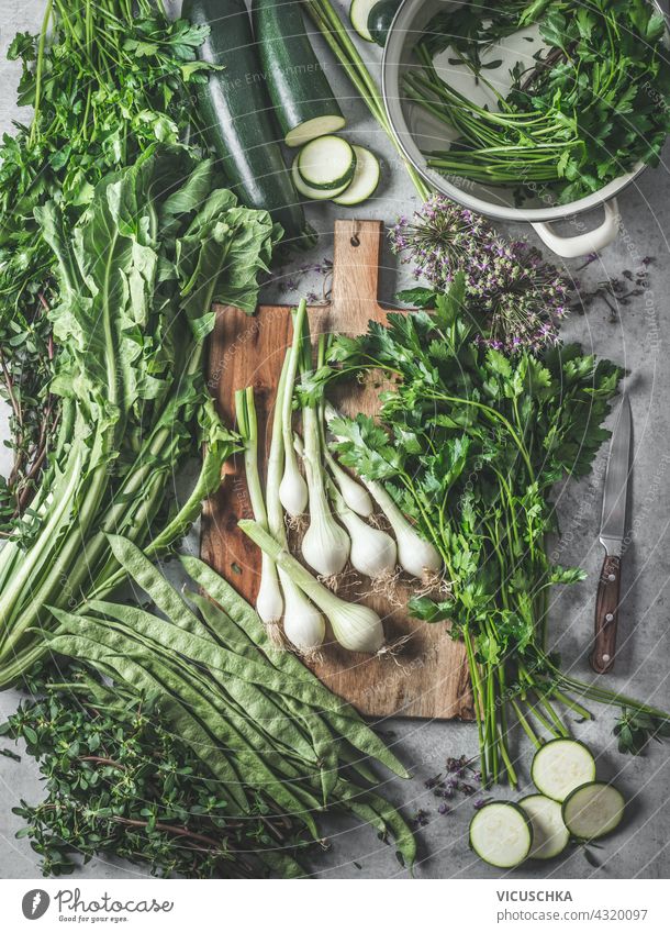 Eine Vielzahl von grünem Gemüse und Kräutern auf einem rustikalen Küchentisch mit einem hölzernen Schneidebrett und Pfannen für gesundes Kochen zu Hause. Ansicht von oben.