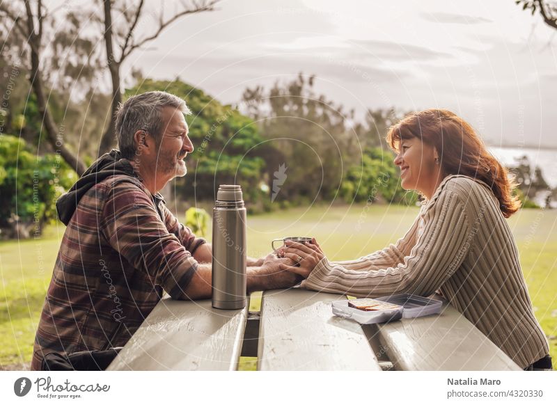 Paar mittleren Alters sitzt auf den Bänken mit Tisch im Park und trinkt Tee. Frau Bank Mann Glück Meer Zusammensein Freizeit Liebe Erwachsener hübsch