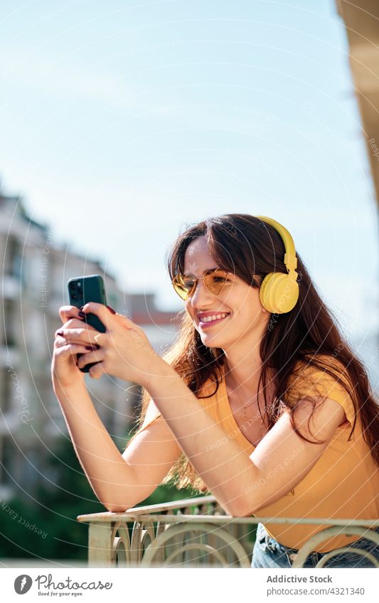 Glückliche Frau mit Kopfhörern, die auf dem Balkon Musik hört zuhören Smartphone benutzend genießen Gesang Melodie Apparatur Gerät Mobile Sommer Browsen