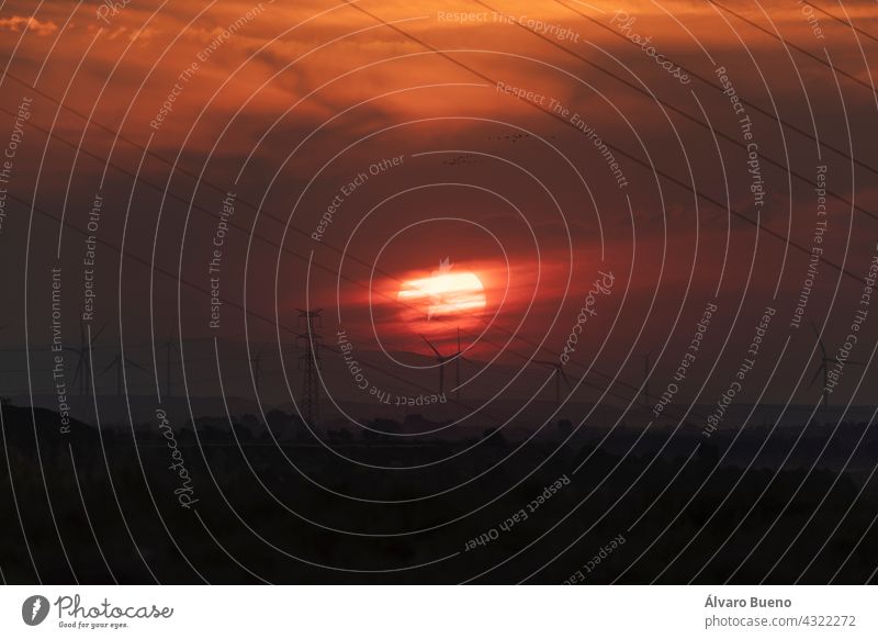 Eine riesige rote Sonne geht hinter den Wolken und einer Landschaft aus Kabeln und Windturbinen im Ebro-Tal unter, in der Nähe der Stadt Gallur, Aragon, Spanien