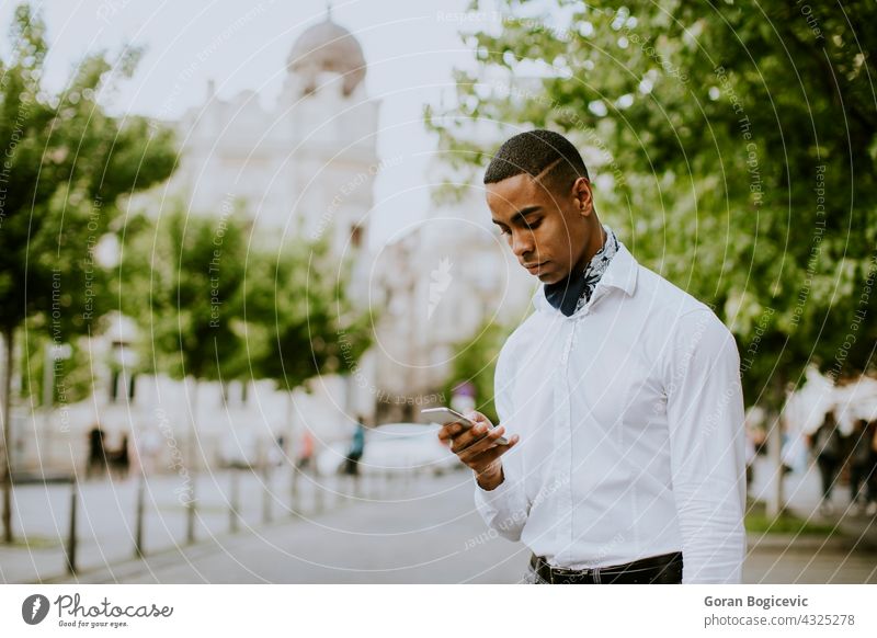 Junger afroamerikanischer Geschäftsmann, der ein Mobiltelefon benutzt, während er auf einer Straße auf ein Taxi wartet Amerikaner schwarz Gebäude Business