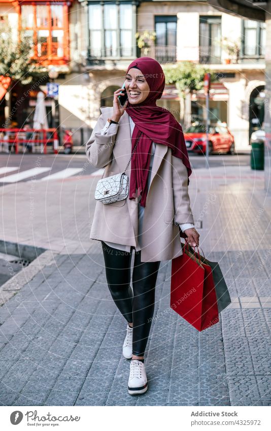 Lächelnde muslimische Frau mit Einkaufstüten in der Stadt reden Smartphone Einkaufstasche Tüte Käufer Großstadt Mobile Anruf Hijab ethnisch Apparatur sprechen