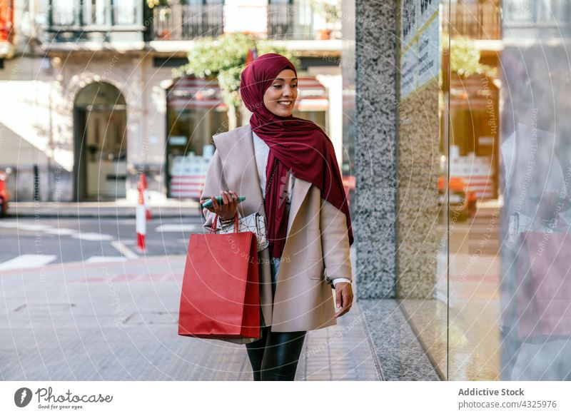 Lächelnde muslimische Frau mit Einkaufstüten in der Nähe eines Schaufensters Käufer Vitrine Großstadt Einkaufstasche Laden Werkstatt konsumgeil Kunde ethnisch