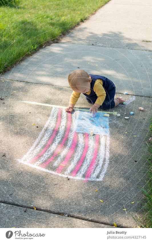 Ein Jahr altes Baby neben der Kreidezeichnung der amerikanischen Flagge; Kleinkind krabbelt und spielt mit Kreide am 4. Juli Stars and Stripes Fahne USA