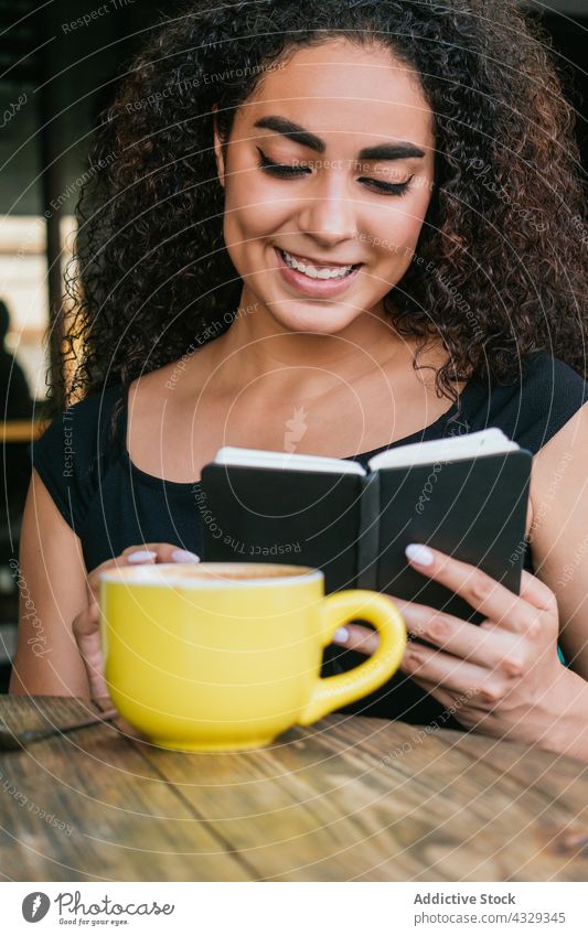 Glückliche Frau liest in einem Café in einem Notizbuch lesen Notebook Kaffee Lächeln Tasse genießen Kaffeepause jung ethnisch hispanisch krause Haare Terrasse