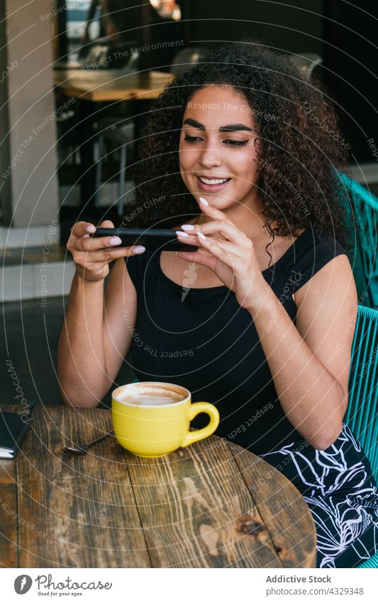 Ethnische Frau benutzt Smartphone in einem Café im Freien Kaffee benutzend fotografieren Tasse Glück Freizeit Telefon jung hispanisch ethnisch krause Haare