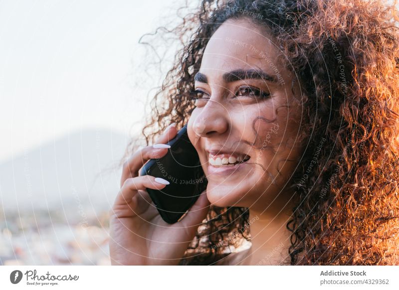Glückliche hispanische Frau, die mit einem Smartphone spricht reden Lächeln krause Haare Telefon Kommunizieren heiter Gespräch jung ethnisch