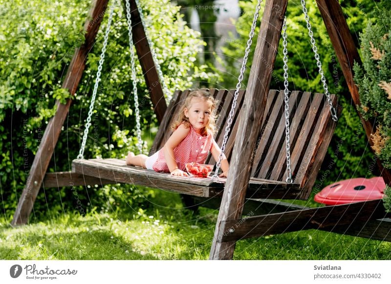Ein kleines Mädchen liegt auf einer Gartenschaukel und isst frische Erdbeeren Baby Essen pendeln sich hinlegen Kleinkind Sommer niedlich Spaß wenig Frucht