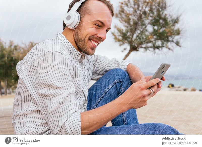 Glücklicher Mann mit Kopfhörern und Smartphone am Strand benutzend zuhören Apparatur Telefon Musik männlich heiter jung Stil Sommer Drahtlos Gerät genießen