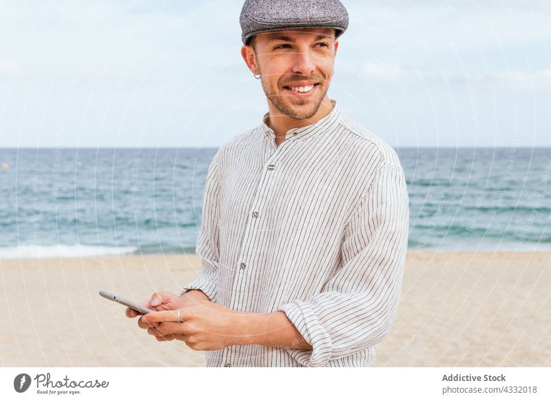 Gut gelaunter Mann mit Smartphone am Sandstrand Strand benutzend Stil Glück MEER trendy Lächeln Telefon männlich jung Vollbart Aussehen Outfit Mode positiv