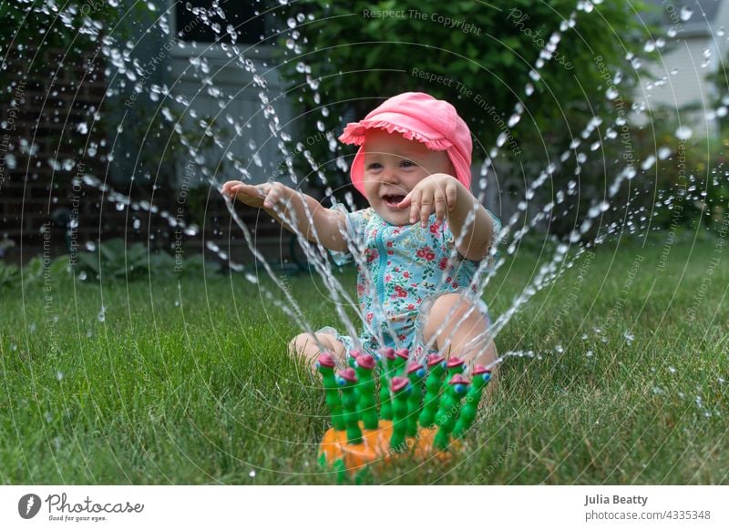 Ein Jahr altes Baby spielt fröhlich im Rasensprenger; einfache Wasserspiel-Unterhaltung für Kleinkind Sonnensicherheit Sprinkleranlage Hof heimwärts Viertel