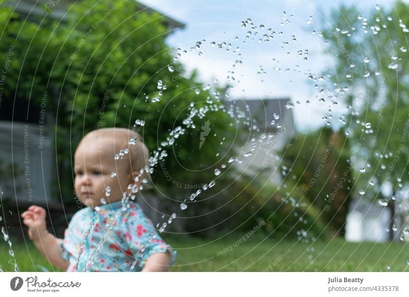 Kleinkind im Gras sitzend im Vorgarten; vor ihr sprüht ein Sprinkler Wasser Baby Sonnensicherheit Sprinkleranlage Wasserspiel Hof heimwärts Viertel