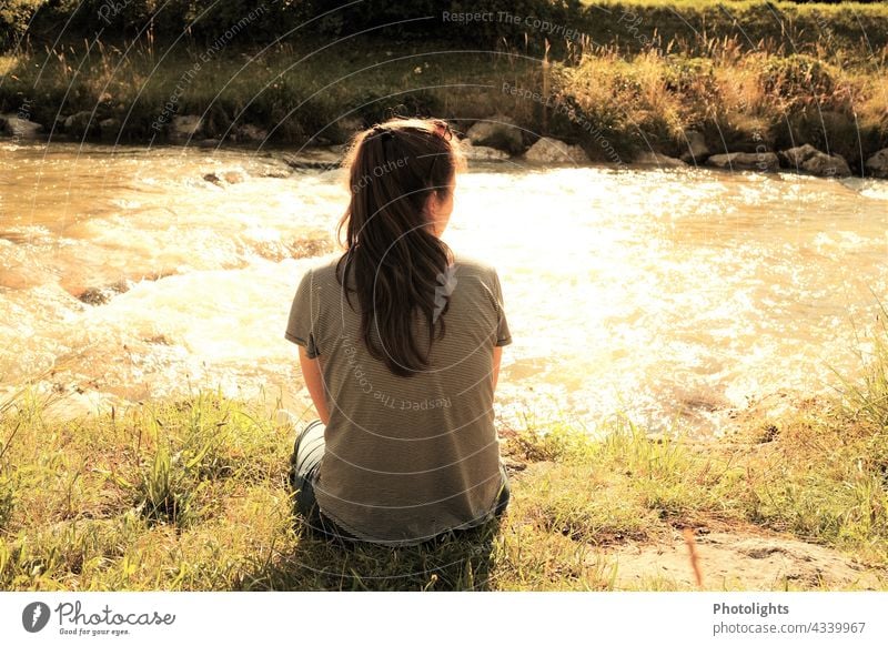 Junge Frau sitzt an einem Bach in der Abendsonne. uferzone Wasser Fluss langhaarig Leben Gelassenheit träumen Schönes Wetter Licht Außenaufnahme Farbfoto