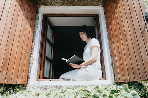 Eine junge Frau mit einem Buch sitzt auf der Fensterbank, Lesen und Entspannen während eines sonnigen Tages Teenager Person Bildung im Innenbereich lesen Raum
