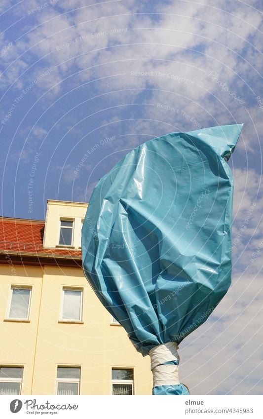mit einem blauen Plastiksack verhülltes Verkehrszeichen vor einem Mehrfamilienhaus im Sonnenschein mit Dekowolken / Baustelle / ungültiges Verkehrszeichen