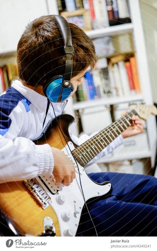 Kind, das zu Hause E-Gitarre spielt, mit einem Kopfhörer und einer unscharfen Bibliothek im Hintergrund, selektiver Fokus elektrisch Stromgitarre Junge Kinder