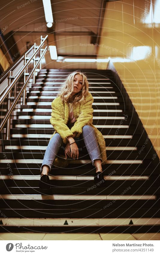 Porträt einer blonden kaukasischen Frau auf der Treppe eines Bahnhofs Mode Lifestyle lässig Mantel gelb Stil jung attraktiv im Innenbereich selbstbewusst urban
