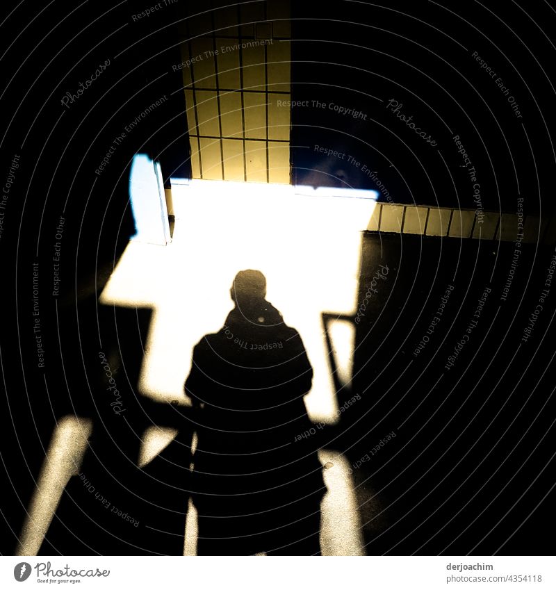 Der Fotograf als schwarzer großer Schatten, tritt in das Treppenhaus das vor ihm von hellem Sonnenlicht durchflutet ist. Vor ihm und rechts sind an der Wand gelbe Fliesen angebracht.