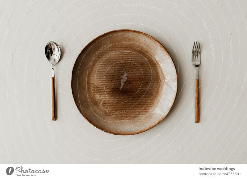 Ein brauner Teller mit einem Löffel und einer Gabel auf beigem Hintergrund. Flachlage, Ansicht von oben. Braune und naturfarbene Teller. Texturiertes körniges Muster auf den Tellern.