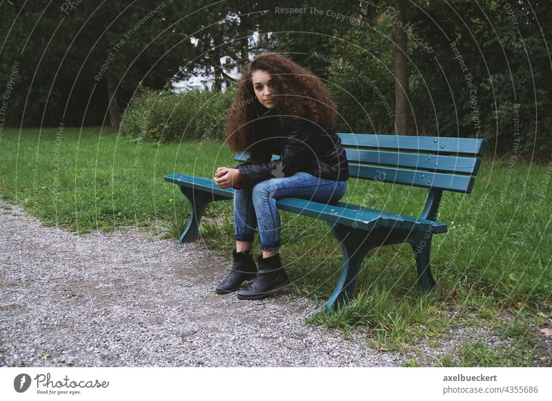 junge Frau sitzt auf Parkbank Junge Frau teenager sitzen warten brünett Locken Jeanshose düster Einsamkeit allein Blick in die Kamera Jugendliche langhaarig Tag