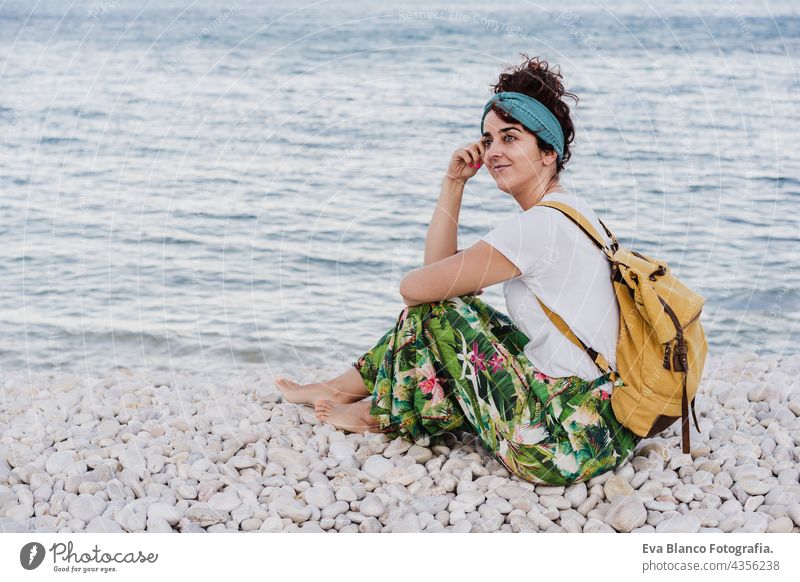 Seitenansicht der entspannten kaukasischen Frau mit gelben Rucksack sitzen am Strand während des Sonnenuntergangs. Sommerzeit. Tagträumen. Lebensstil im Freien