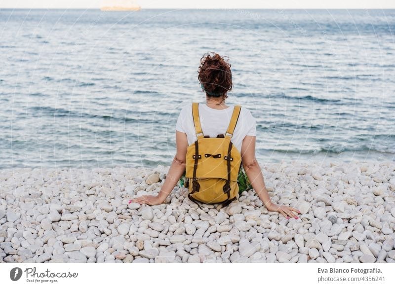 Rückenansicht der entspannten kaukasischen Frau mit gelben Rucksack sitzen am Strand während des Sonnenuntergangs. Sommerzeit. Lebensstil im Freien Kaukasierin