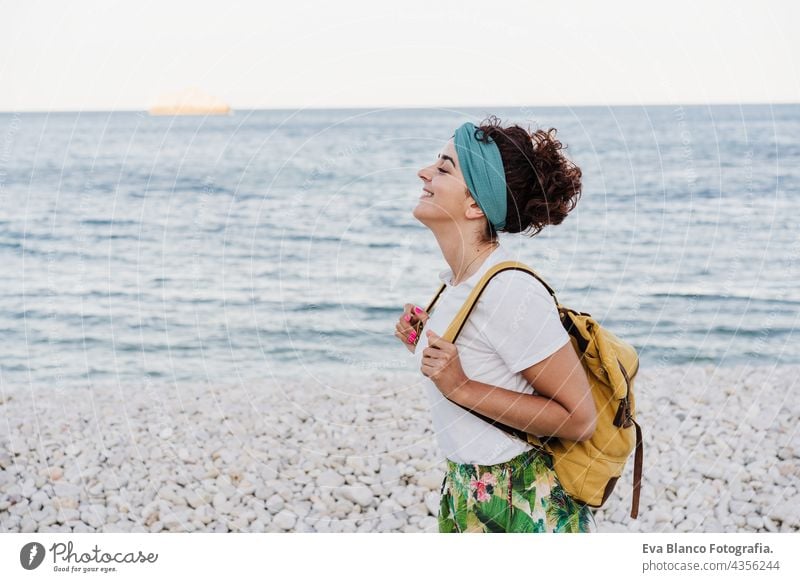entspannte kaukasische Frau mit gelbem Rucksack, die während des Sonnenuntergangs am Strand spazieren geht. Sommerzeit. tagträumen. Lebensstil im Freien