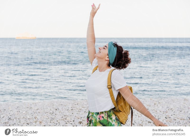glückliche kaukasische Frau mit gelbem Rucksack und erhobenen Armen am Strand bei Sonnenuntergang. Sommerzeit. Tagträumen. Lebensstil im Freien Kaukasierin