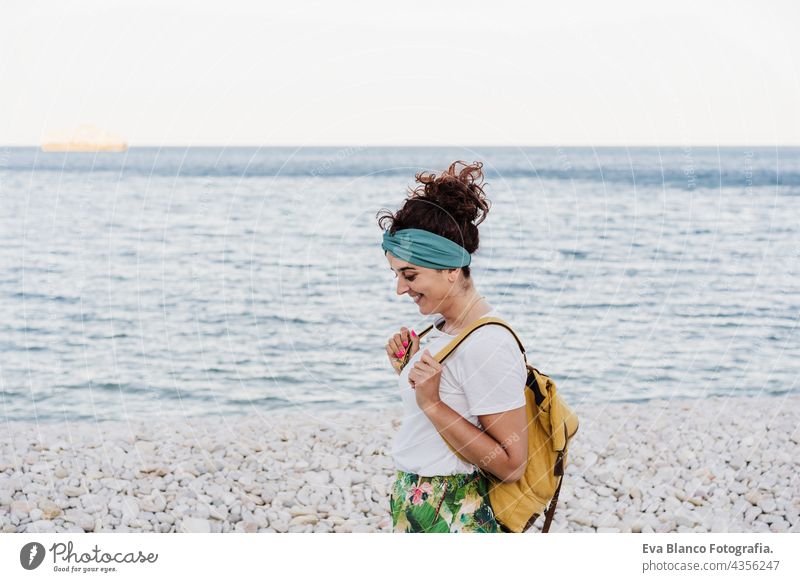 entspannte kaukasische Frau mit gelbem Rucksack, die während des Sonnenuntergangs am Strand spazieren geht. Sommerzeit. tagträumen. Lebensstil im Freien