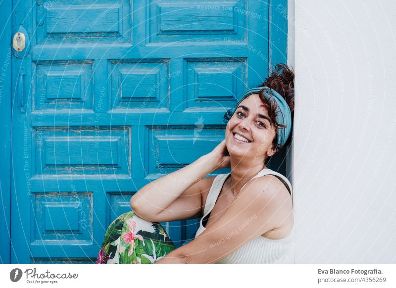 lächelnde kaukasische Frau, die im Sommer vor einer blauen Tür sitzt. Lebensstil im Freien Glück Kaukasier blaue Tür Großstadt Strand sorgenfrei Lächeln