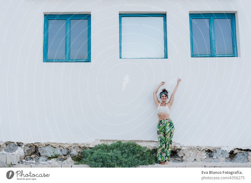 Porträt einer glücklichen kaukasischen Frau mit erhobenen Armen vor blauen Fenstern und weißer Wand im Sommer. Lebensstil im Freien Glück Kaukasier Großstadt