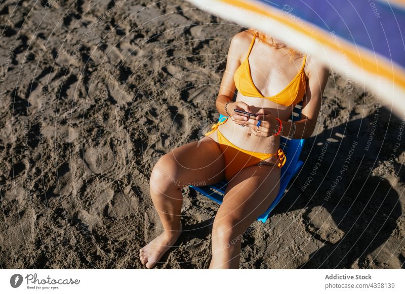 Anonyme Frau mit Telefon am Strand sitzend Bikini benutzend passen Sommer Urlaub Sonnenschirm MEER reisen Technik & Technologie Browsen Feiertag blass anonym
