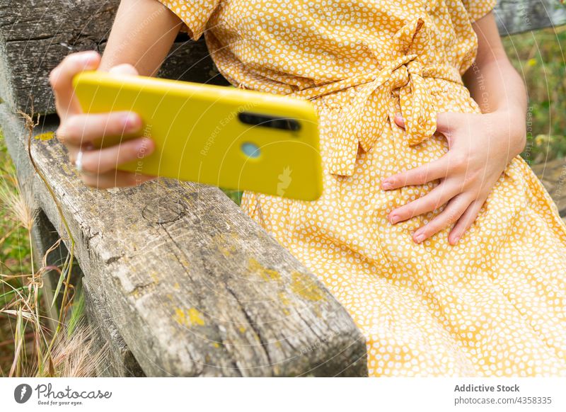 Anonyme schwangere Frau, die ein Selfie mit ihrem Smartphone auf dem Land macht Selbstportrait berühren Bauch Sommer Landschaft sitzen ruhen Kleid Mutter