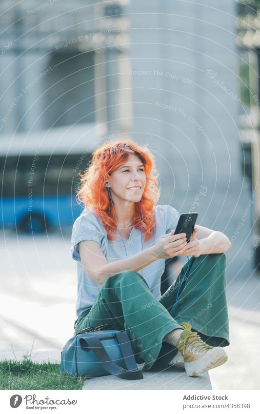 Vergnügte Frau mit roten Haaren, die auf der Straße über Kopfhörer Musik hört zuhören Smartphone Browsen Rotschopf Nachricht soziale Netzwerke rote Haare Gesang