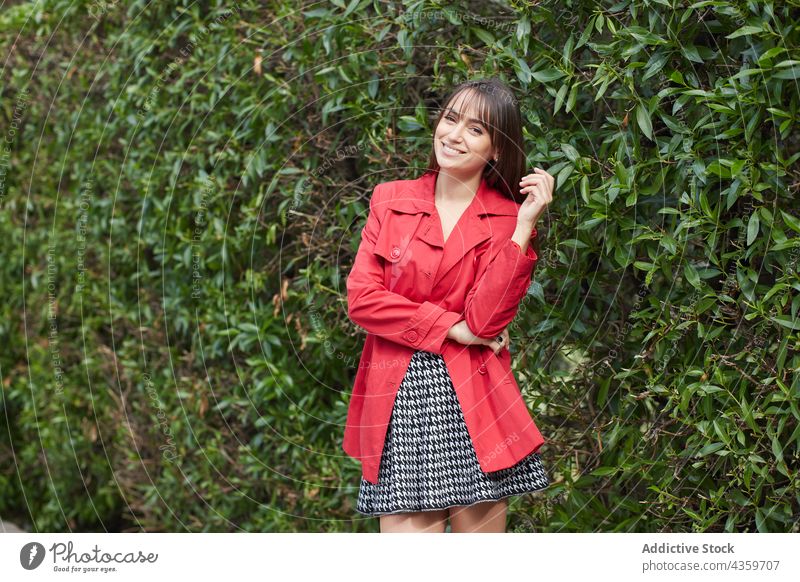 Lächelnde junge Frau in roter Jacke im Park stehend Stil Mode trendy Frühling Outfit Oberbekleidung Farbe Stilrichtung Glück heiter Bekleidung anhaben brünett