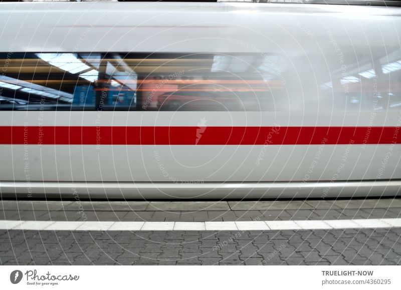 Ein schneeweisser ICE Zug mit einem dicken roten Streifen verlässt gerade München Hauptbahnhof. In seinen Fenstern spiegelt sich die Bahnhofshalle mit Neonleuchten, das graue Pflaster des Bahnsteigs zeigt einen weißen Streifen