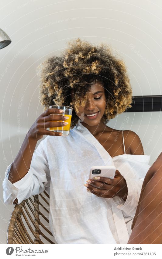 Glückliche schwarze Frau sitzt auf einem Sessel und surft auf ihrem Smartphone benutzend Internet Lächeln online Texten Nachricht trinken zuschauen digital