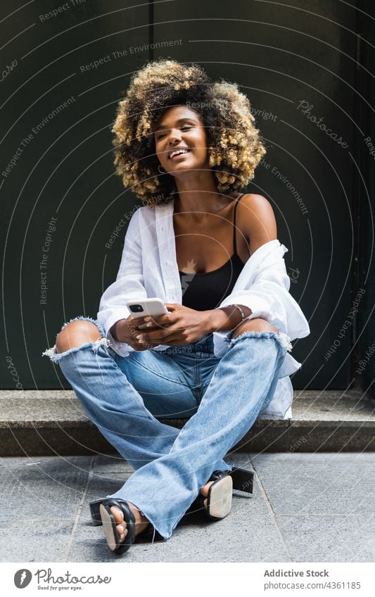 Lächelnde schwarze Frau, die ihr Smartphone benutzt, während sie auf einer Betongrenze sitzt positiv Internet benutzend genießen Surfen digital Glück angenehm