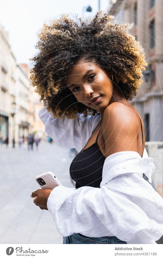 Schwarze Frau berührt ihr Haar, während sie mit ihrem Smartphone auf der Straße steht Individualität cool ernst urban tausendjährig Laufsteg Körperhaltung schön