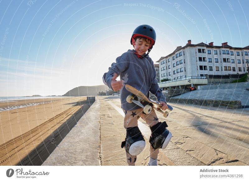 Lächelnder Teenager-Skater mit Skateboard auf einer Böschung stehend Junge Stauanlage Meeresufer Sommer MEER Aktivität genießen heiter Glück sitzen Spazierweg
