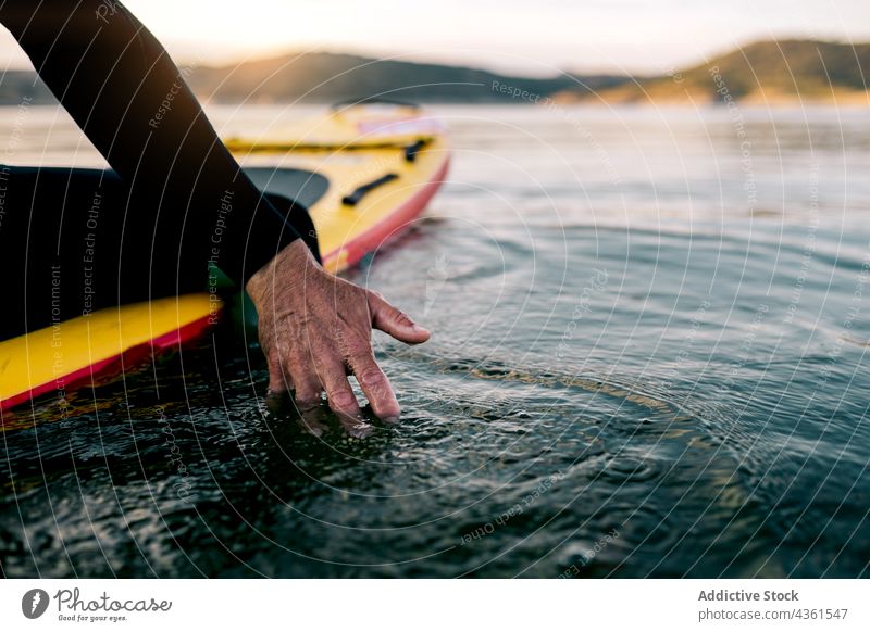 Crop Mann auf Paddleboard berühren Meerwasser Paddelbrett Zusatzplatine MEER Wasser Surfer Sonnenuntergang Sommer friedlich männlich Abend Rippeln Oberfläche