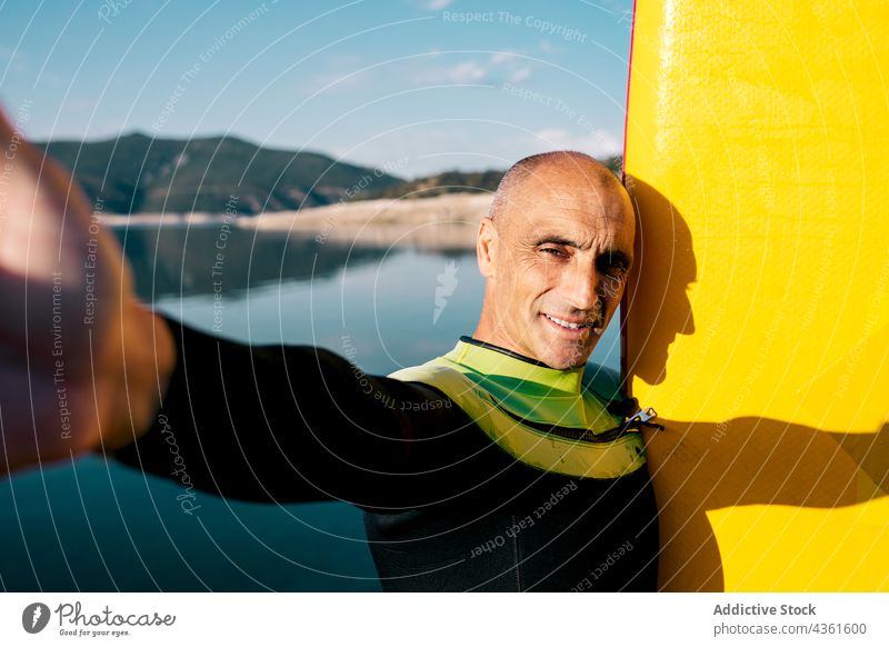 Lächelnder reifer Mann mit Paddelbrett, der ein Selfie am Seeufer macht Holzplatte Aktivität üben sportlich gelb männlich Lebensmitte Wassersport Hobby aktiv