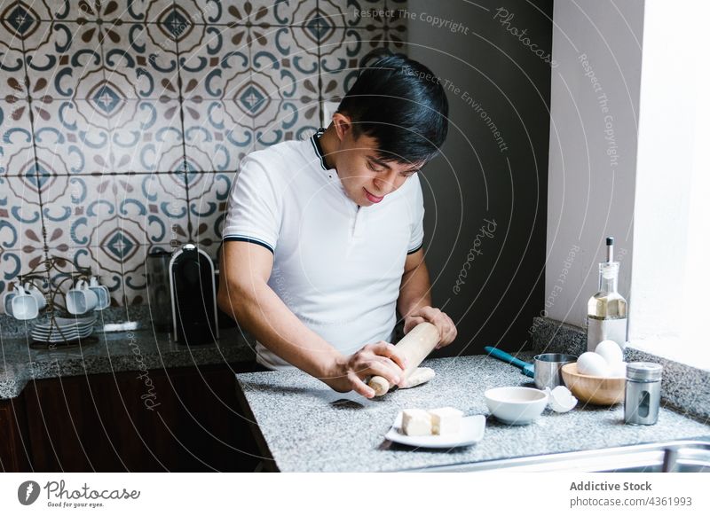 Lateinischer Junge mit Down-Syndrom rollt Teig am Tisch Koch rollen Teigwaren Teenager vorbereiten Gebäck Küche ethnisch Lächeln Glück heimwärts Lebensmittel