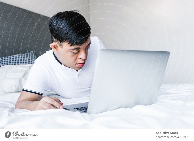 Ethnischer Teenager mit Down-Syndrom surft zu Hause auf seinem Laptop Junge Browsen Lügen Bett Surfen benutzend heimwärts ethnisch Latein unterhalten Apparatur