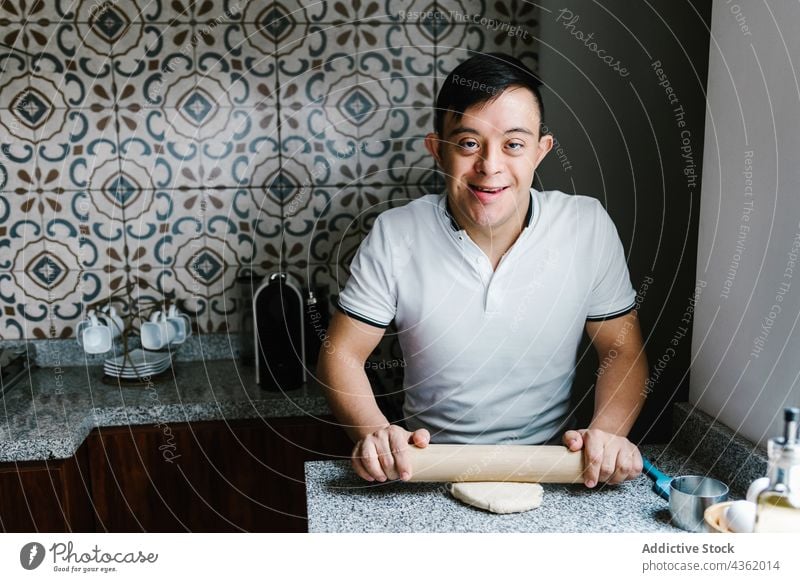Glücklicher lateinischer Junge mit Down-Syndrom rollt Teig am Tisch Koch rollen Teigwaren heiter Teenager vorbereiten Gebäck Küche ethnisch Latein Lächeln