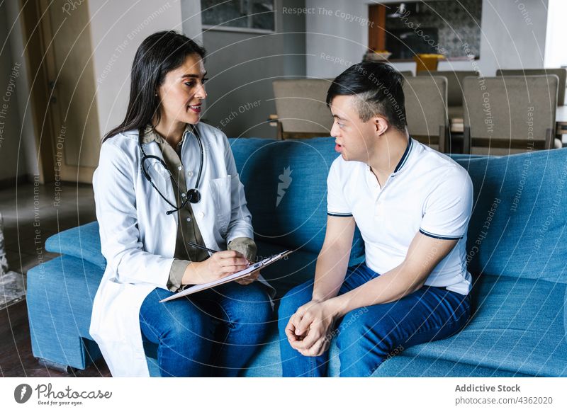 Zufriedener Arzt im Gespräch mit einem lateinamerikanischen Jungen mit Down-Syndrom bei einem Besuch zu Hause geduldig Ernennung reden zur Kenntnis nehmen