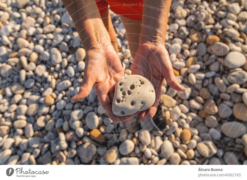 Eine Frau in den 70ern hält einen Stein voller Löcher an einem natürlichen Strand am Mittelmeer, Spanien Textur Objekt Steinstrand Hände Nahaufnahme Senior
