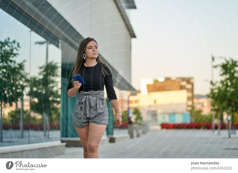 junge Frau geht mit Handy in der Hand neben einem Büro Telefon Smartphone klug Zelle Funktelefon laufen Spaziergang gehen Sie Dame Frauen Uhr hispanisch