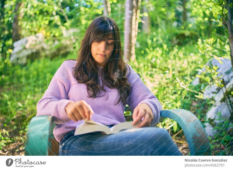 Entspannte Latina-Frau liest ein Buch auf einem türkisfarbenen Vintage-Stuhl, draußen auf ihrer Veranda, umgeben von Natur, Bäumen und Gras. lateinamerikanisch