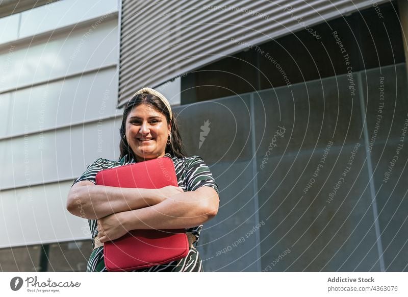 Glückliche Frau mit Laptop-Tasche in der Nähe eines städtischen Gebäudes Lächeln Fall urban modern kurvenreich Stil Farbe Übergröße Streifen Geometrie jung